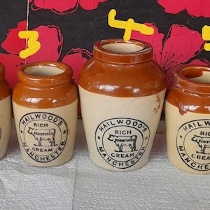 Puede incluir: Seis tarros de crema antiguos con tapas y bases marrones y cuerpos blanquecinos. Cada tarro presenta el texto "HAILWOOD'S RICH CREAM MANCHESTER" y un gráfico de vaca. Los tarros están dispuestos en fila.