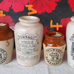 Op de afbeelding: Een verzameling van zes antieke roompotten. De potten zijn in verschillende maten en vormen, met een crème- en bruin kleurenschema. Sommige potten hebben etiketten met tekst en illustraties, waaronder "Fresh Cream".