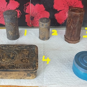 May include: A collection of five items: two small cylindrical metal containers, one tall hexagonal brown container, one rectangular metal box with an embossed floral and circular design, and one round blue plastic container with a lid.