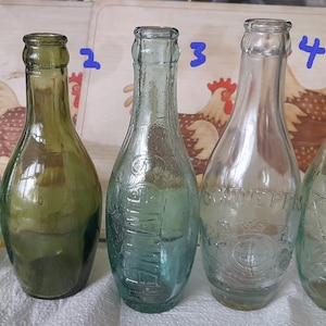 Puede incluir: Se muestran cinco botellas de vidrio antiguas de varios colores y formas. Las botellas son transparentes, verdes y verde azulado, con diseños y marcas en relieve. El fondo presenta una tabla de cortar de madera con un diseño de gallo.