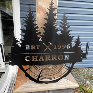 May include: Black metal sign with a silhouette of pine trees, a saw blade, and the text "EST 1996 CHARRON".