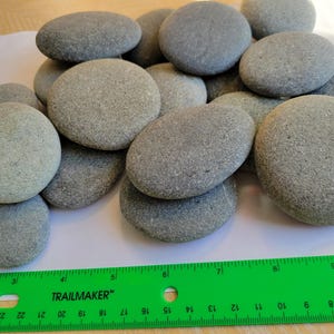 May include: A collection of smooth, rounded gray river stones of varying sizes arranged in a pile. A green plastic ruler is positioned at the bottom, showing the stones range from approximately 1 to 3 inches in diameter.