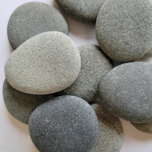 May include: A collection of smooth, rounded river stones in various shades of gray, ranging from light slate to dark charcoal. The stones are irregularly shaped, featuring textured, matte surfaces and are clustered together in a dense, overlapping arrangement.