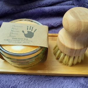 May include: A glass jar containing a natural dish soap bar, a wooden brush with natural bristles, and a bamboo tray. The jar has a label that reads "Handmade Artisan Naturally Derived Soaps Dish Soap Bar No plastic or chemicals, strong cleansing Made on the Wirral - with love"