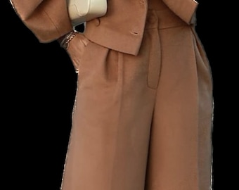 May include: A tan two-piece suit featuring a short jacket with large sleeves and matching wide-leg pants. A small, cream-colored purse hangs from the arm. The outfit is accessorized with a silver watch and rings.