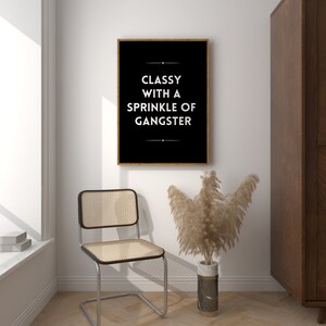 May include: A framed black and white print with the text "Classy with a Sprinkle of Gangster" hangs on a white wall. A chrome chair with a woven seat and back sits below the print. A vase with dried plants is next to the chair.
