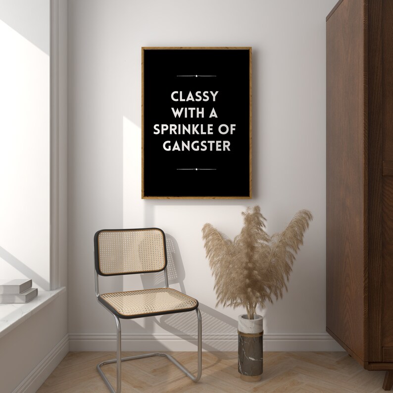 May include: A framed black and white print with the text "Classy with a Sprinkle of Gangster" hangs on a white wall. A chrome chair with a woven seat and back sits below the print. A vase with dried plants is next to the chair.