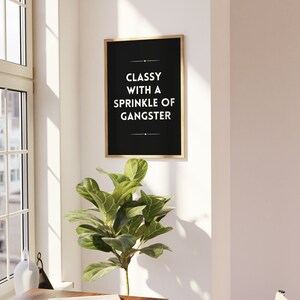 May include: A framed black print with gold trim hangs on a white wall. The print reads "Classy with a Sprinkle of Gangster" in white text. A green leafy plant sits below the print, near a window.