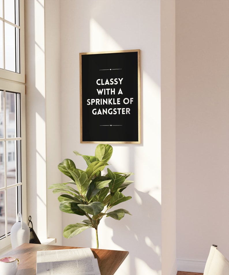 May include: A framed black print with gold trim hangs on a white wall. The print reads "Classy with a Sprinkle of Gangster" in white text. A green leafy plant sits below the print, near a window.