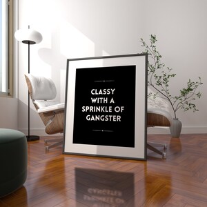 May include: A framed black and white print with the text "Classy with a Sprinkle of Gangster." The print is leaning against a wall in a room with a modern chair, floor lamp, and decorative plant. The room has a hardwood floor.