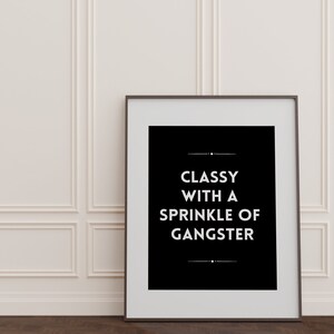 May include: A framed black and white print with the text "CLASSY WITH A SPRINKLE OF GANGSTER" against a white wall. The frame is dark, and the wall has decorative molding. The floor is dark wood.