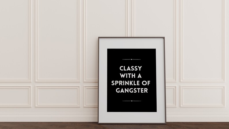 May include: A framed black and white print with the text "CLASSY WITH A SPRINKLE OF GANGSTER" against a white wall. The frame is dark, and the wall has decorative molding. The floor is dark wood.