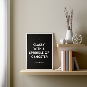 May include: A framed black and white print with the text "Classy with a Sprinkle of Gangster" displayed on a wooden shelf. The shelf also holds books, decorative objects, and a white vase with branches.