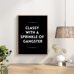 May include: A framed black and white print with the text "CLASSY WITH A SPRINKLE OF GANGSTER." The print is in a light brown wooden frame. The image is on a white wall, with a wooden table, a plant, and a hanging light fixture.