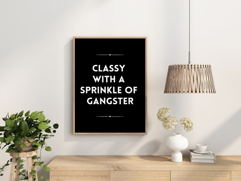May include: A framed black and white print with the text "CLASSY WITH A SPRINKLE OF GANGSTER." The print is in a light brown wooden frame. The image is on a white wall, with a wooden table, a plant, and a hanging light fixture.