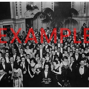 May include: A black and white photograph of a large group of people gathered in a ballroom. The people are dressed in formal attire and are smiling and laughing. The photo was taken at the Canadian Hotel on July 24th, 1921.
