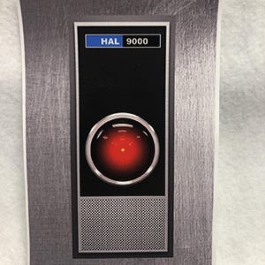 May include: A rectangular image featuring a design inspired by the HAL 9000 computer from the movie 2001: A Space Odyssey. The design includes a red glowing eye, a speaker grill, and the text "HAL 9000" in a blue box.