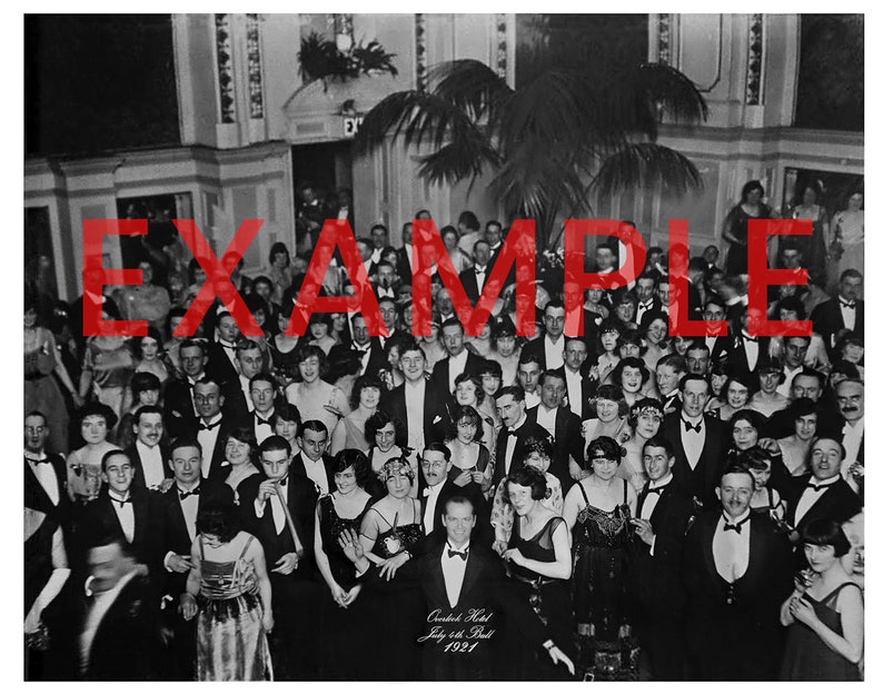 May include: A black and white photograph of a large group of people gathered in a ballroom. The people are dressed in formal attire and are smiling and laughing. The photo was taken at the Canadian Hotel on July 24th, 1921.