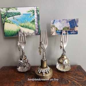 May include: Three unique fork-shaped card holders with decorative bases. Each holds a small landscape painting. The forks are adorned with charms and sit on ornate bases. The holders are displayed on a wooden surface.