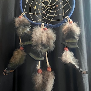 May include: A handcrafted dreamcatcher with a navy blue hoop and a woven web design. The dreamcatcher features gray feathers, wooden beads, and a decorative stone at the top. This item is designed for home decor.