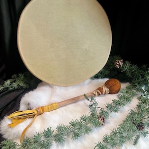 May include: A large, round drum with a light tan drumhead, positioned above a wooden drum beater with a brown head and yellow leather accents. The drum and beater rest on a white, fluffy surface, surrounded by green sprigs.