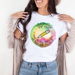 May include: White t-shirt with a colorful watercolor circle design featuring a yellow pencil and the text "You've prepared your best. Your testing strategies will help with the rest!"