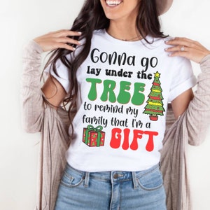 May include: White t-shirt with a green Christmas tree and red gift box graphic. The text reads "Gonna go lay under the TREE to remind my family that I'm a GIFT".