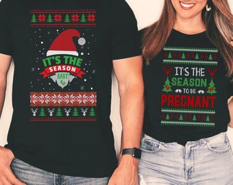 May include: Black t-shirts with Christmas-themed designs. One shirt features a Santa hat and the text "IT'S THE SEASON BABY!" with a baby graphic. The other shirt says "IT'S THE SEASON TO BE PREGNANT" with a Christmas tree and deer.