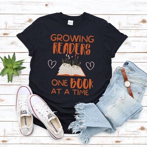 Może przedstawiać: Czarny T-shirt z napisem "Growing Readers One Book At A Time" w kolorze pomarańczowym. Na koszulce znajduje się stylizowany otwarty książka z kwiatami rosnącymi z niej i dwoma sercami.