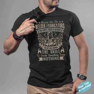 May include: Black t-shirt with a graphic design featuring a welder and the text "Blessed Are The Welder Fabricators." The design includes wings, stars, and the phrase "The Skill To Create Something From Nothing."