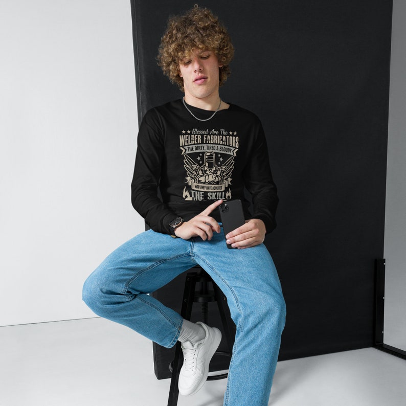 May include: Black long-sleeve shirt with a graphic that reads "Blessed Are The Welder Fabricators The Dirty, Tired & Bloody For They Have Acquired The Skill." The shirt is worn with blue jeans and white sneakers.