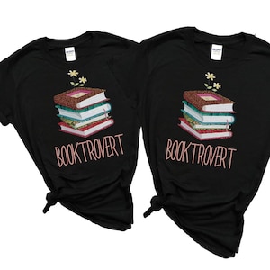 Puede incluir: Dos camisetas negras con una pila de libros y flores en la parte superior. El texto "BOOKTROVERT" está impreso en rosa en cada camiseta.