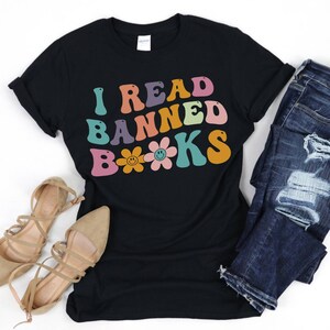 Peut inclure: T-shirt noir avec le texte "I Read Banned Books" en couleurs arc-en-ciel avec des fleurs à smiley.