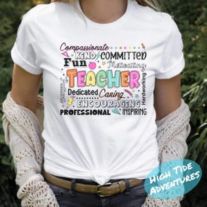 May include: White t-shirt with the word "TEACHER" in colorful letters. Surrounding the word are other positive adjectives like "Compassionate," "Kind," "Dedicated," and "Inspiring." The shirt has a casual, everyday style.