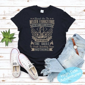 May include: Black t-shirt with a graphic featuring a welder and the text "Blessed Are The Welder Fabricators." The design includes wings, stars, and the phrase "The Skill To Create Something From Nothing." White sneakers and blue jeans are also in the image.