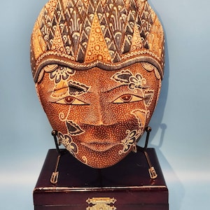 May include: A brown wooden mask with intricate patterns and floral designs. The mask features a serene face with detailed eyes and a decorative headpiece. It is displayed on a black metal stand atop a dark wooden box with a brass clasp.