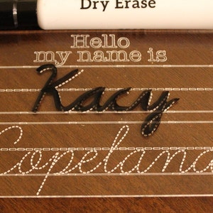 以下が含まれることがあります： 練習用の筆記体を書くための点線が入った透明なプラスチック製のドライイレースボードです。ボードには「Hello my name is」と黒のマーカーで書かれた「Kacy Copeland」の名前が書かれています。