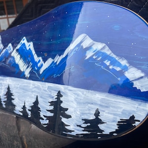 May include: A skateboard deck featuring a hand-painted night landscape with snow-capped blue mountains, a dark blue sky with white stars, a foreground of black pine tree silhouettes, and a partial red and yellow circular emblem on the left side.