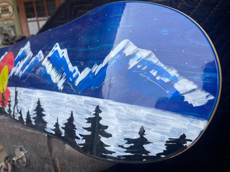 May include: A skateboard deck featuring a hand-painted night landscape with snow-capped blue mountains, a dark blue sky with white stars, a foreground of black pine tree silhouettes, and a partial red and yellow circular emblem on the left side.
