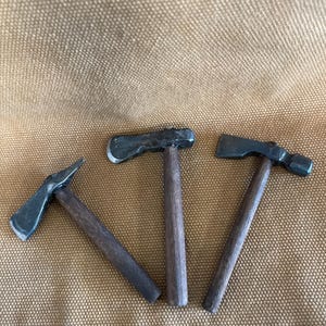 Könnte beinhalten: Drei Miniaturwerkzeuge mit dunklen Metallköpfen und Holzgriffen. Das Set enthält ein pickelartiges Werkzeug und zwei kleine Äxte, die jeweils eine texturierte, dunkelgraue Metalloberfläche und einen geraden, zylindrischen braunen Holzgriff haben.
