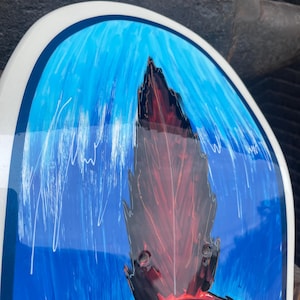May include: A skateboard deck featuring a glossy finish with a vibrant blue background and white vertical brushstroke details. A central dark red and black jagged, flame-like graphic element extends upward, framed by a smooth, rounded white border.