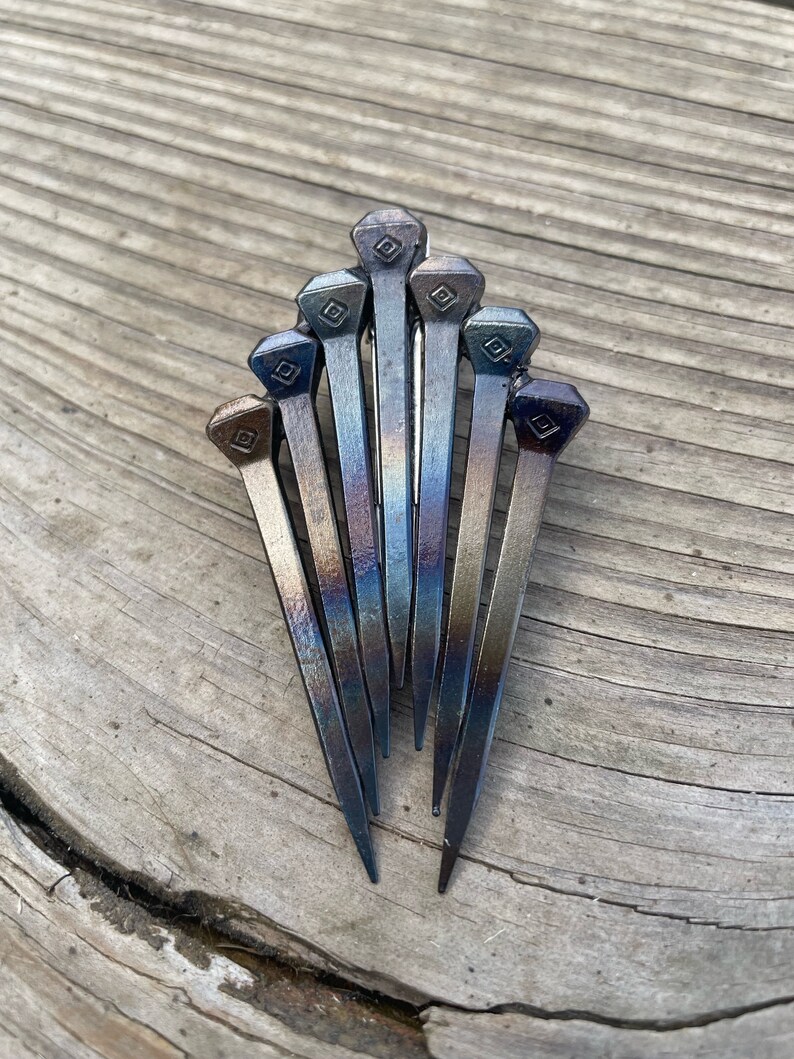 Op de afbeelding: Een verzameling van zeven metalen spijkers gerangschikt in een waaier. De spijkers hebben een donkere, iriserende afwerking met ruitvormige details op de koppen. Ze worden gepresenteerd op een verweerd houten oppervlak.