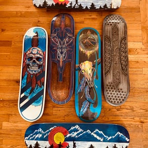May include: Six skateboard decks featuring diverse designs: two with Colorado flag mountain landscapes, one with a skull wearing a headdress, one with blue pinstriping on wood, one with a bull skull and feathers, and one with a grey honeycomb pattern.
