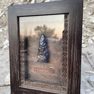 May include: A dark brown wooden frame containing a dark blue, flaked stone arrowhead mounted on a dark wood backing. Below the arrowhead is a small, rectangular copper-colored plaque with the embossed text "APOBKO". The frame features a geometric border.