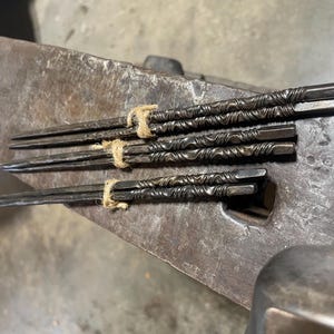以下が含まれることがあります： テクスチャ加工された、ねじれたデザインの、手作りの金属製箸6膳セット。 箸は紐で束ねられ、風化した金属の表面に置かれています。 各箸は約20cmで、素朴な美しさを表現しています。