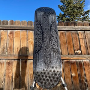 May include: A black skateboard deck featuring a silver glitter finish and a geometric pattern of hexagons and repeating cubes. The design includes a central vertical panel with a textured surface and a pointed top, set against a dark background.