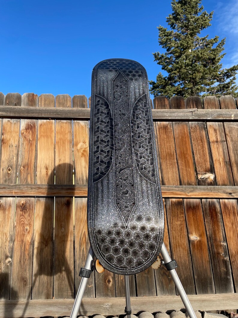May include: A black skateboard deck featuring a silver glitter finish and a geometric pattern of hexagons and repeating cubes. The design includes a central vertical panel with a textured surface and a pointed top, set against a dark background.