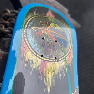 May include: A skateboard deck featuring a bright blue border and a central circular graphic. The design includes a stylised bow and arrows set against a dark background with dripping paint effects in yellow, pink, and white, along with four mounting holes.