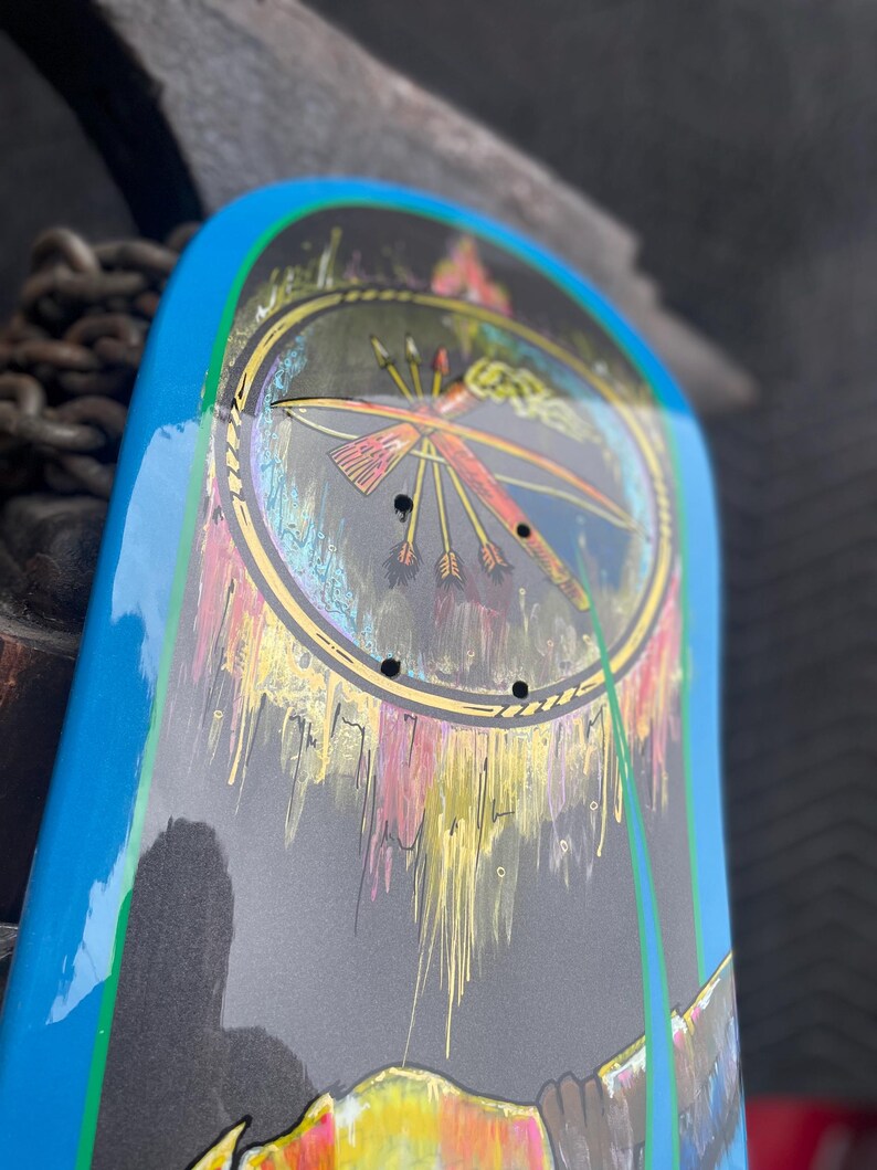 May include: A skateboard deck featuring a bright blue border and a central circular graphic. The design includes a stylised bow and arrows set against a dark background with dripping paint effects in yellow, pink, and white, along with four mounting holes.