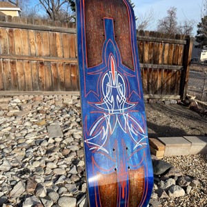May include: A skateboard deck featuring a dark wood grain finish with a blue metallic overlay. The surface is decorated with intricate orange and white pinstripe designs in a symmetrical pattern, including thin lines and pointed shapes.